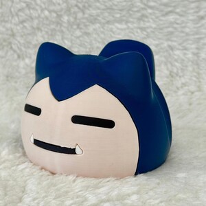 May include: A blue and light pink Snorlax character figurine. The figurine features a light pink face with black eyes and mouth, and a single tooth. The top of the figurine is blue.
