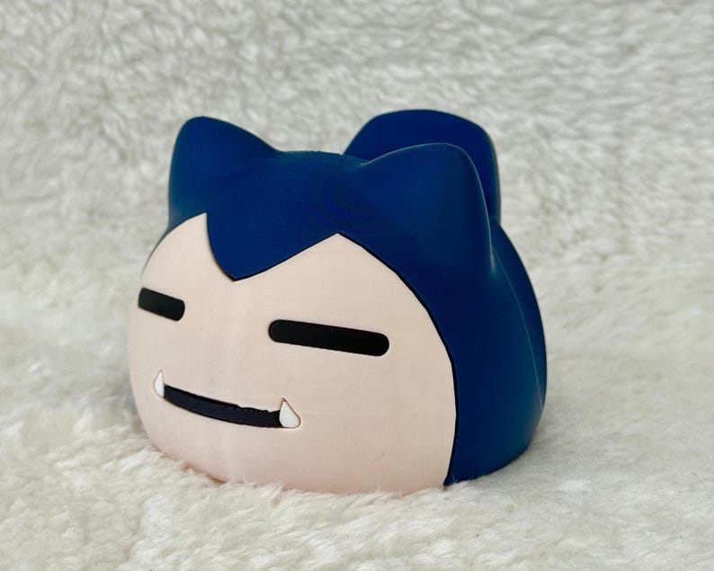 May include: A blue and light pink Snorlax character figurine. The figurine features a light pink face with black eyes and mouth, and a single tooth. The top of the figurine is blue.