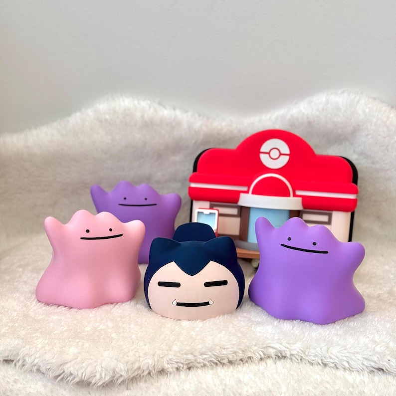 May include: A collection of colorful, cartoon-style figures, including a pink Ditto, a blue Snorlax, and two purple Dittos, are arranged in front of a red and white building. The figures have simple, smiling faces. The scene is set on a white, textured surface.
