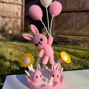 May include: A pink bunny figurine holding three balloons, two pink and one white. The bunny is standing in a pink basket with two eggs, two bunny heads, and yellow and white flowers. The figurine is on a white surface.