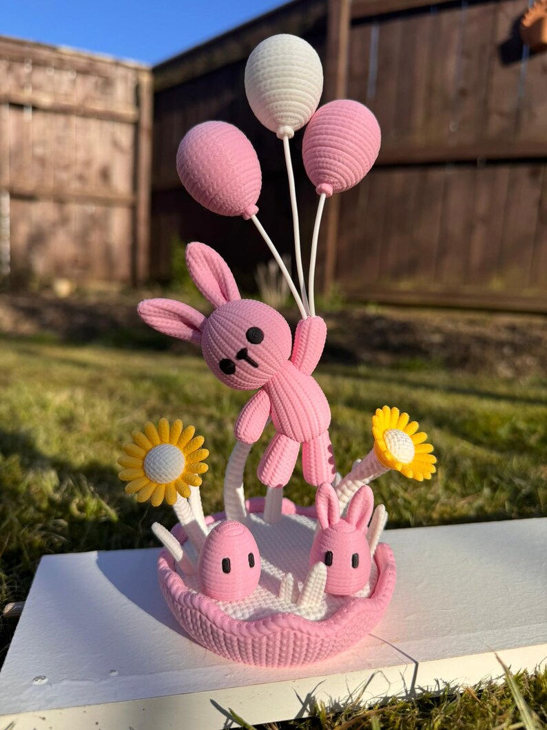 May include: A pink bunny figurine holding three balloons, two pink and one white. The bunny is standing in a pink basket with two eggs, two bunny heads, and yellow and white flowers. The figurine is on a white surface.