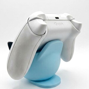 Puede incluir: Un mando Xbox blanco descansa sobre un soporte azul claro. El soporte tiene forma redondeada con una base plana. El logotipo de Xbox es visible en el mando. El fondo es blanco liso.