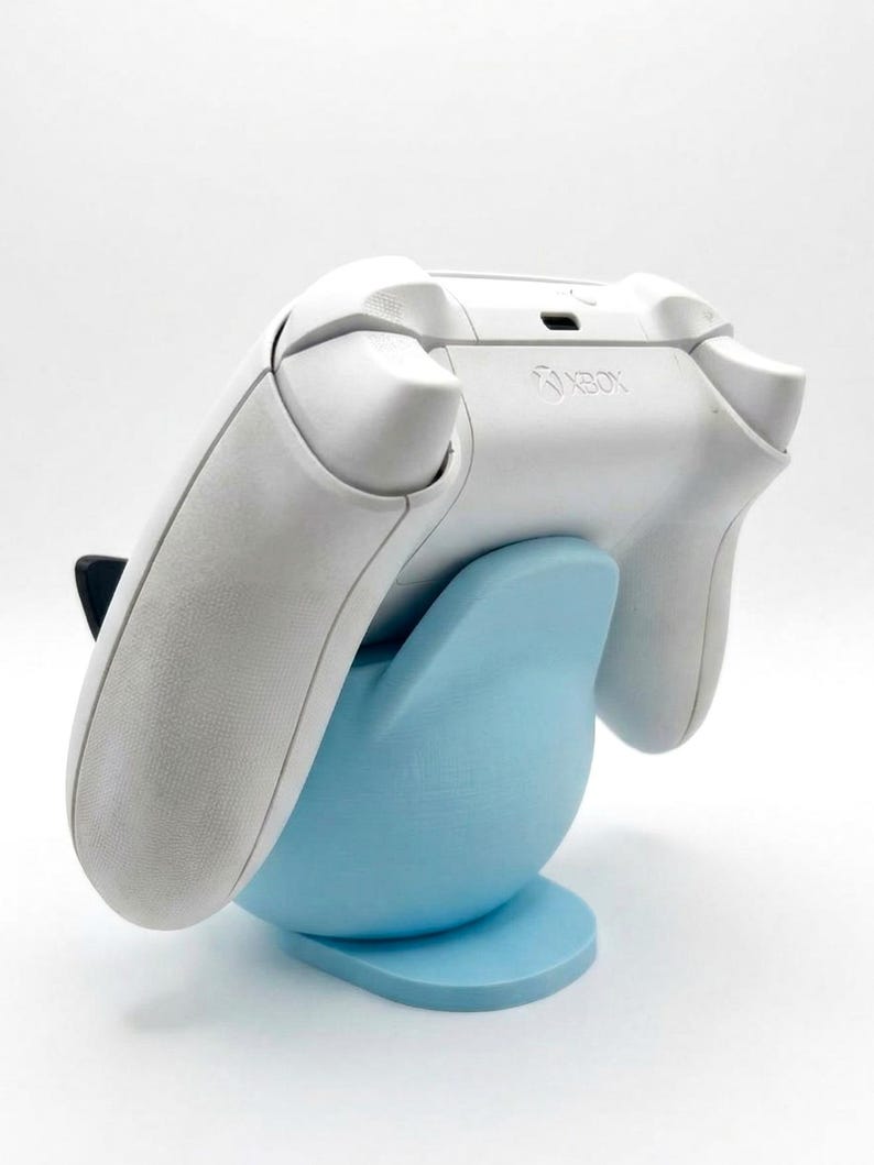 Puede incluir: Un mando Xbox blanco descansa sobre un soporte azul claro. El soporte tiene forma redondeada con una base plana. El logotipo de Xbox es visible en el mando. El fondo es blanco liso.