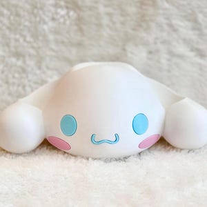 Puede incluir: Un soporte de pl&aacute;stico blanco con forma de personaje que presenta grandes orejas ca&iacute;das, dos ojos azules redondos, una boca azul peque&ntilde;a y dos acentos circulares rosas en las mejillas. El objeto tiene un acabado mate liso y dise&ntilde;o sim&eacute;trico.