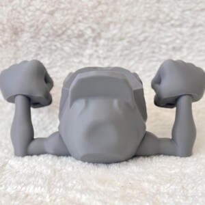May include: A gray, stylized figurine with arms raised in a flexing pose. The figurine has a head and two arms with clenched fists. The object is made of a smooth, solid material and is set against a white background.
