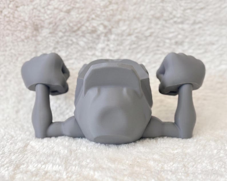 May include: A gray, stylized figurine with arms raised in a flexing pose. The figurine has a head and two arms with clenched fists. The object is made of a smooth, solid material and is set against a white background.