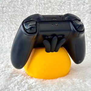 Puede incluir: Un mando de videojuegos negro apoyado sobre un soporte amarillo brillante con forma de mano de tres dedos. El mando cuenta con un puerto micro-USB en la parte superior y un peque&ntilde;o bot&oacute;n circular, con la palabra venom impresa en el centro del soporte.