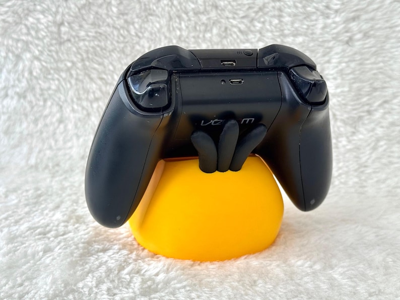 Puede incluir: Un mando de videojuegos negro apoyado sobre un soporte amarillo brillante con forma de mano de tres dedos. El mando cuenta con un puerto micro-USB en la parte superior y un peque&ntilde;o bot&oacute;n circular, con la palabra venom impresa en el centro del soporte.
