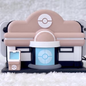 May include: A beige and white gaming console dock shaped like a building with a central blue door featuring a circular emblem. The structure includes a small matching kiosk on the left side and a black base, designed to hold a handheld gaming device.