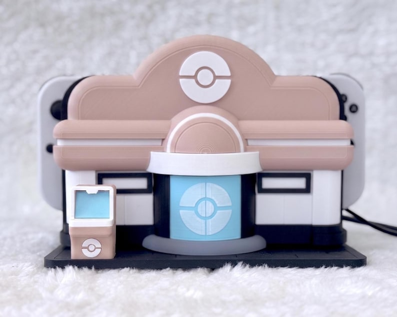 May include: A beige and white gaming console dock shaped like a building with a central blue door featuring a circular emblem. The structure includes a small matching kiosk on the left side and a black base, designed to hold a handheld gaming device.