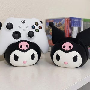 May include: A white Xbox wireless controller resting on a black and white Kuromi-themed character stand, positioned next to an identical empty Kuromi character stand. Both stands feature a pink skull emblem on the forehead and pointed ears.