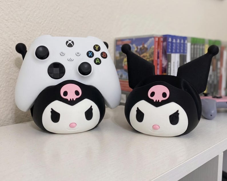 May include: A white Xbox wireless controller resting on a black and white Kuromi-themed character stand, positioned next to an identical empty Kuromi character stand. Both stands feature a pink skull emblem on the forehead and pointed ears.
