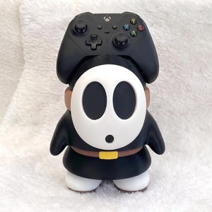 May include: A black video game controller resting on top of a Shy Guy character figure. The figure has a white oval mask with black eye and mouth holes, a black robe, a brown belt with a yellow buckle, and white shoes. The controller is black.