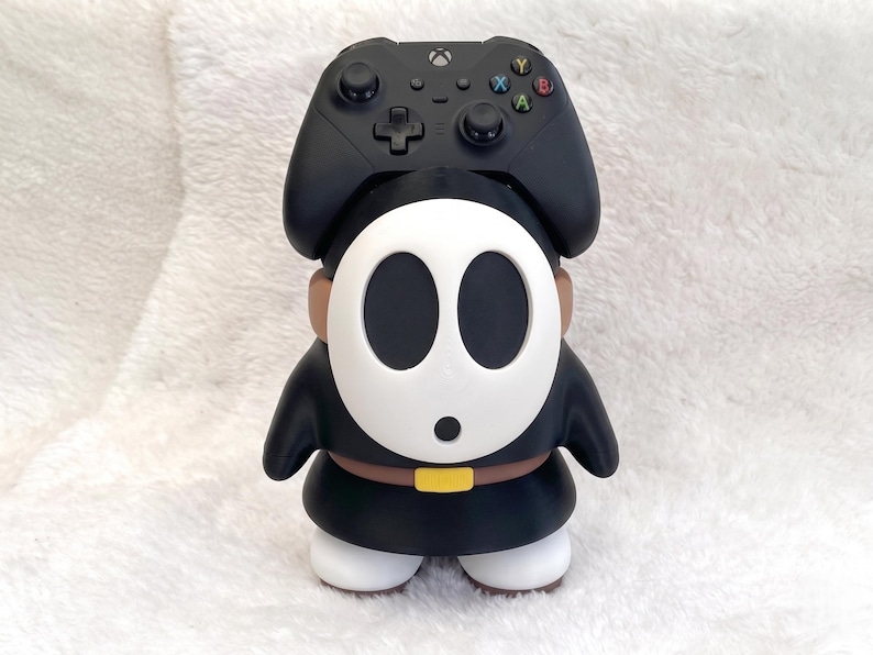 May include: A black video game controller resting on top of a Shy Guy character figure. The figure has a white oval mask with black eye and mouth holes, a black robe, a brown belt with a yellow buckle, and white shoes. The controller is black.