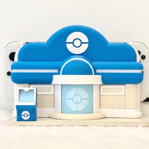 May include: A blue and white plastic gaming console dock shaped like a building with a circular emblem on the front. The structure features a central entrance, a small side kiosk, and a flat base, designed to hold a handheld gaming device.