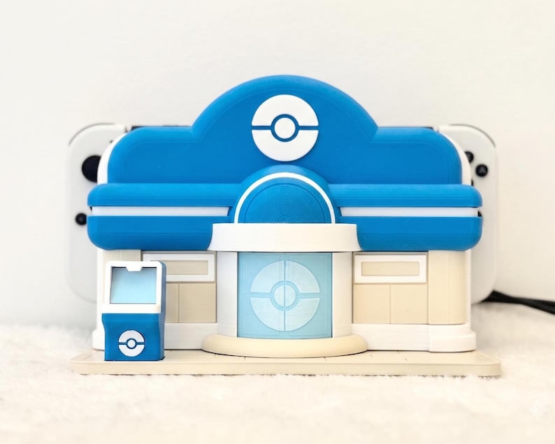 May include: A blue and white plastic gaming console dock shaped like a building with a circular emblem on the front. The structure features a central entrance, a small side kiosk, and a flat base, designed to hold a handheld gaming device.