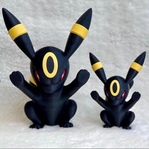 Puede incluir: Dos figuras de Umbreon negras y amarillas con ojos rojos. La figura m&aacute;s grande mide 23 cm de altura, mientras que la m&aacute;s peque&ntilde;a mide 15 cm. Ambas figuras presentan orejas puntiagudas con bandas amarillas y una marca ovalada amarilla en la frente.