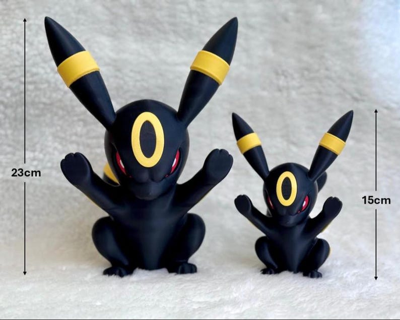 Puede incluir: Dos figuras de Umbreon negras y amarillas con ojos rojos. La figura m&aacute;s grande mide 23 cm de altura, mientras que la m&aacute;s peque&ntilde;a mide 15 cm. Ambas figuras presentan orejas puntiagudas con bandas amarillas y una marca ovalada amarilla en la frente.