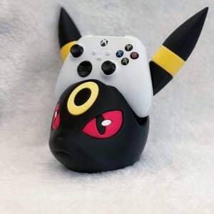 May include: A black and yellow character-themed controller stand featuring pointed ears and red eyes, holding a white wireless gaming controller with black joysticks and multicolored action buttons on its top surface.