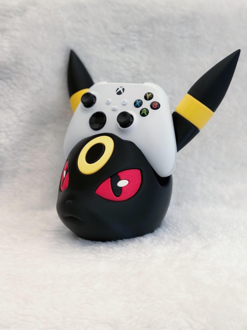 May include: A black and yellow character-themed controller stand featuring pointed ears and red eyes, holding a white wireless gaming controller with black joysticks and multicolored action buttons on its top surface.