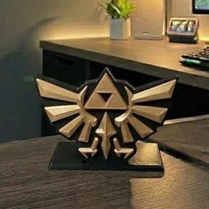 May include: A black and gold decorative emblem featuring a stylized design with a triangle and wings. The emblem is on a black base and appears to be a 3D-printed item, likely a collectible or decorative piece.