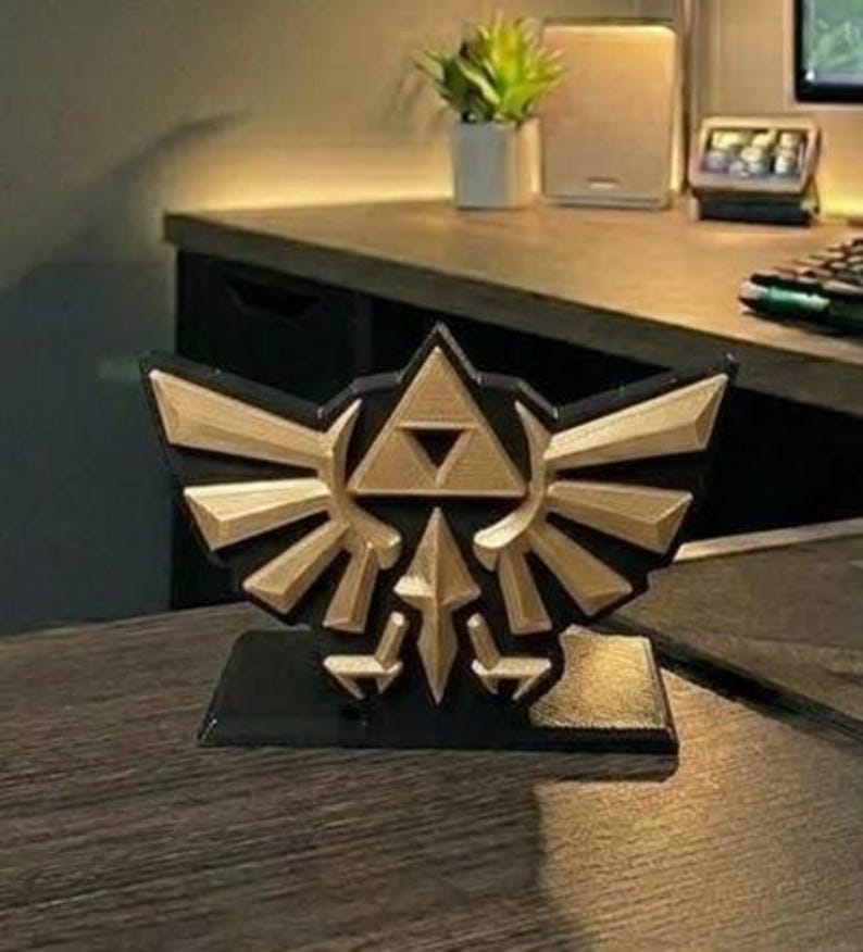 May include: A black and gold decorative emblem featuring a stylized design with a triangle and wings. The emblem is on a black base and appears to be a 3D-printed item, likely a collectible or decorative piece.