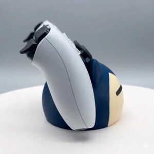 May include: A white and black games controller sits in a blue and tan character-themed holder. The controller is angled, showing the buttons and triggers. The holder has a rounded shape with a cartoon-like design.