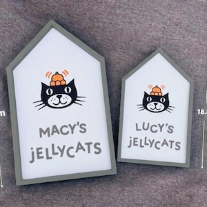 May include: Two house-shaped signs with a gray border and white interior. Each sign features a black cat illustration wearing a hat, with the text "MACY'S JELLYCATS" and "LUCY'S JELLYCATS". The larger sign is 26.2 cm tall, and the smaller one is 18.8 cm tall.