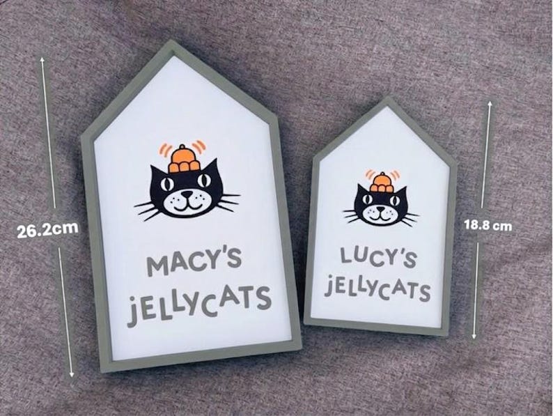 May include: Two house-shaped signs with a gray border and white interior. Each sign features a black cat illustration wearing a hat, with the text "MACY'S JELLYCATS" and "LUCY'S JELLYCATS". The larger sign is 26.2 cm tall, and the smaller one is 18.8 cm tall.