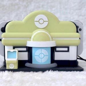 May include: A pale yellow and white gaming console dock shaped like a building with a central blue door, a circular emblem on the roof, and a small matching kiosk to the left, designed to hold a handheld gaming device with white controllers.
