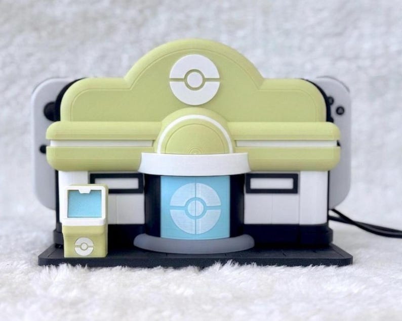 May include: A pale yellow and white gaming console dock shaped like a building with a central blue door, a circular emblem on the roof, and a small matching kiosk to the left, designed to hold a handheld gaming device with white controllers.