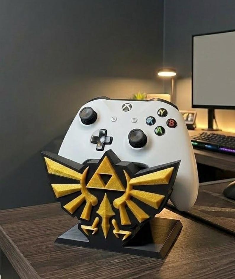 May include: A white Xbox controller sits on a black and gold decorative stand. The stand features a gold emblem with a black base. The controller has black buttons and coloured action buttons.