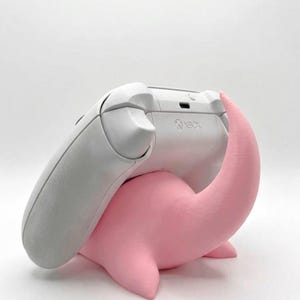 Pode incluir: Um controle Xbox branco repousa sobre um suporte rosa estilizado em forma de Nessie. O controle est&aacute; inclinado, mostrando o logotipo do Xbox. O suporte tem um acabamento liso e fosco, e o controle &eacute; de cor cinza claro.