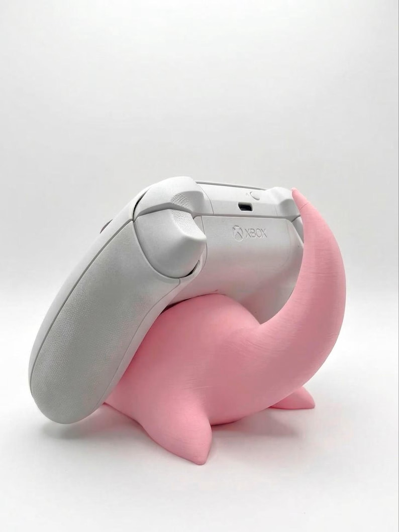 Pode incluir: Um controle Xbox branco repousa sobre um suporte rosa estilizado em forma de Nessie. O controle est&aacute; inclinado, mostrando o logotipo do Xbox. O suporte tem um acabamento liso e fosco, e o controle &eacute; de cor cinza claro.