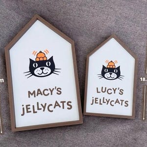 May include: Two house-shaped wooden signs with white backgrounds and brown frames. Each sign features a black cat illustration with an orange hat and the text "MACY'S JELLYCATS" and "LUCY'S JELLYCATS". The larger sign is 26.2 cm tall.