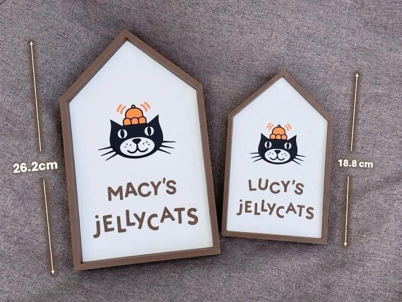 May include: Two house-shaped wooden signs with white backgrounds and brown frames. Each sign features a black cat illustration with an orange hat and the text "MACY'S JELLYCATS" and "LUCY'S JELLYCATS". The larger sign is 26.2 cm tall.