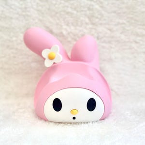 May include: A pink and white plastic character figure featuring a rabbit-like head with long ears, one of which is bent. The face is white with black oval eyes, a small yellow nose, and a tiny black mouth. A white flower with a yellow center is attached.