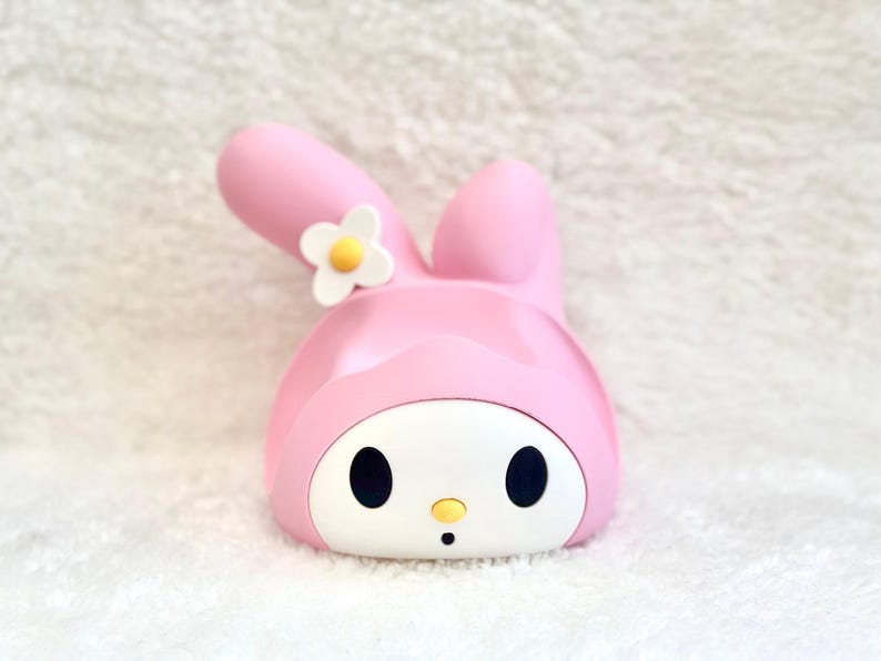 May include: A pink and white plastic character figure featuring a rabbit-like head with long ears, one of which is bent. The face is white with black oval eyes, a small yellow nose, and a tiny black mouth. A white flower with a yellow center is attached.