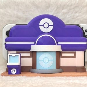 May include: A purple and white 3D-printed model of a building, resembling a Pok&eacute;mon Center. The model has a detailed design with a circular logo on the roof and a light blue central structure. The model is placed in front of a white and black game console.