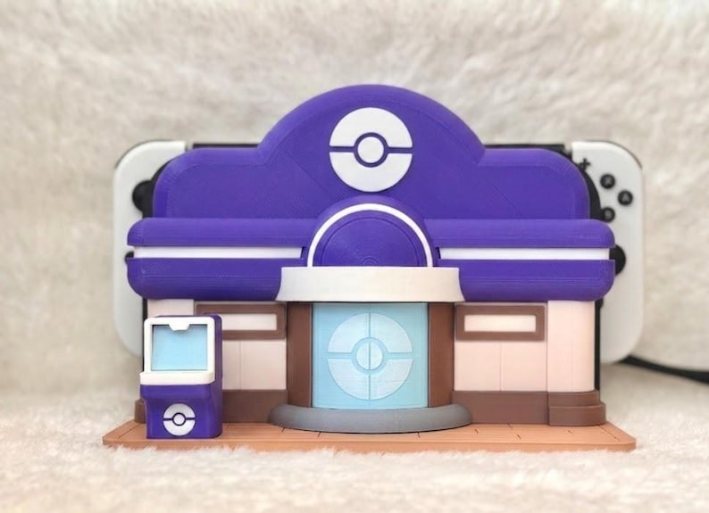 May include: A purple and white 3D-printed model of a building, resembling a Pok&eacute;mon Center. The model has a detailed design with a circular logo on the roof and a light blue central structure. The model is placed in front of a white and black game console.