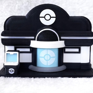 May include: A black and white model of a building featuring a central entrance with a light blue circular emblem, a matching emblem on the roof, and a small standalone kiosk to the left with a light blue screen and a circular logo on the base.