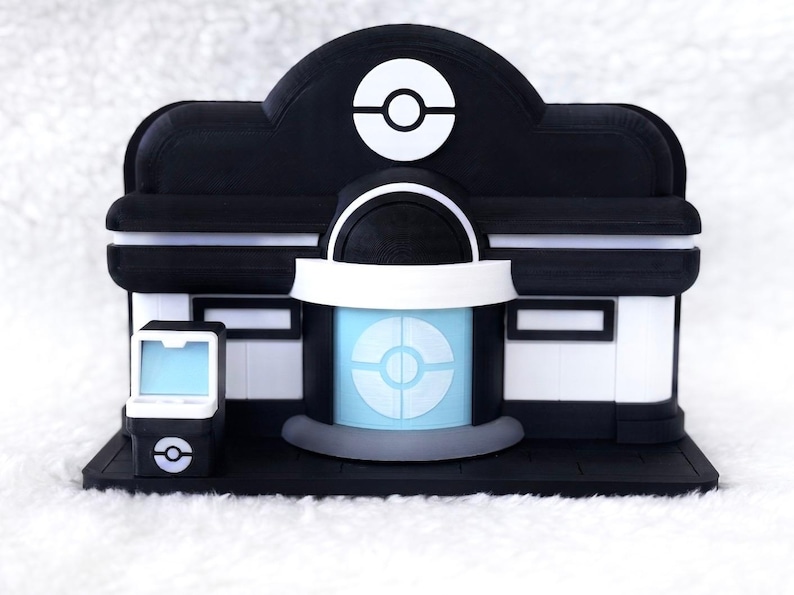 May include: A black and white model of a building featuring a central entrance with a light blue circular emblem, a matching emblem on the roof, and a small standalone kiosk to the left with a light blue screen and a circular logo on the base.