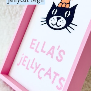 Puede incluir: Un letrero rectangular con marco rosa y fondo blanco que presenta una ilustraci&oacute;n de un gato negro con un sombrero naranja. El texto dice ELLA'S JELLYCATS en letras rosas. Las palabras Jellycat Sign aparecen en un recuadro azul arriba.