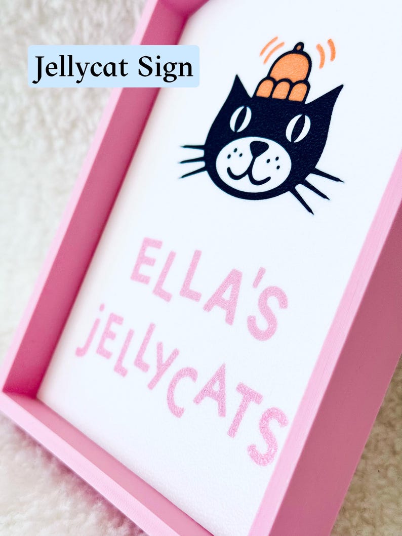 Puede incluir: Un letrero rectangular con marco rosa y fondo blanco que presenta una ilustraci&oacute;n de un gato negro con un sombrero naranja. El texto dice ELLA'S JELLYCATS en letras rosas. Las palabras Jellycat Sign aparecen en un recuadro azul arriba.