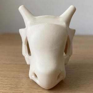 May include: A cream-colored, 3D-printed skull with prominent horns and facial features. The skull has a smooth, matte finish and is set against a light-colored wooden surface. The design is reminiscent of a fantasy creature.