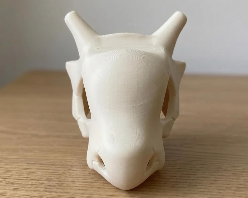 May include: A cream-colored, 3D-printed skull with prominent horns and facial features. The skull has a smooth, matte finish and is set against a light-colored wooden surface. The design is reminiscent of a fantasy creature.