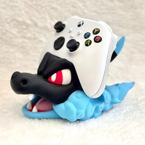May include: A white Xbox wireless controller resting on a black and light blue creature-shaped stand. The stand features a black head with red eyes, a light blue cloud-like base, and an open mouth showing white teeth and a pink tongue.