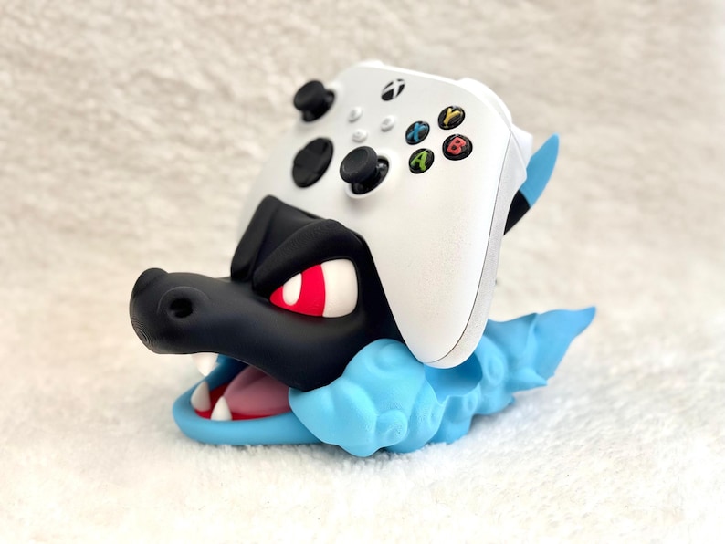May include: A white Xbox wireless controller resting on a black and light blue creature-shaped stand. The stand features a black head with red eyes, a light blue cloud-like base, and an open mouth showing white teeth and a pink tongue.