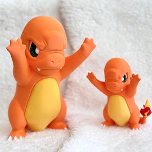May include: Two orange and yellow Charmander figures standing side by side. Both figures have raised arms, green eyes, and white claws. The smaller figure on the right features a red and yellow flame detail on the tip of its tail.