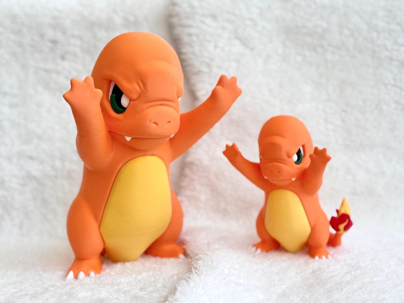 May include: Two orange and yellow Charmander figures standing side by side. Both figures have raised arms, green eyes, and white claws. The smaller figure on the right features a red and yellow flame detail on the tip of its tail.
